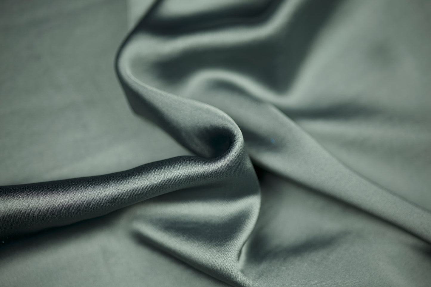 Nahaufnahme eines luxuriösen Satinstoffs in elegantem Salbeigrün mit sanftem Schimmer und natürlichem Faltenwurf – ideal für dezente Mode und Wohntextilien.
